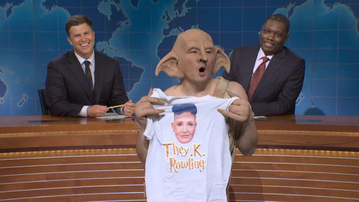 SNL cast member in a costume holding a J.K. Rowling shirt, while hosts smile in a news desk comedy sketch.