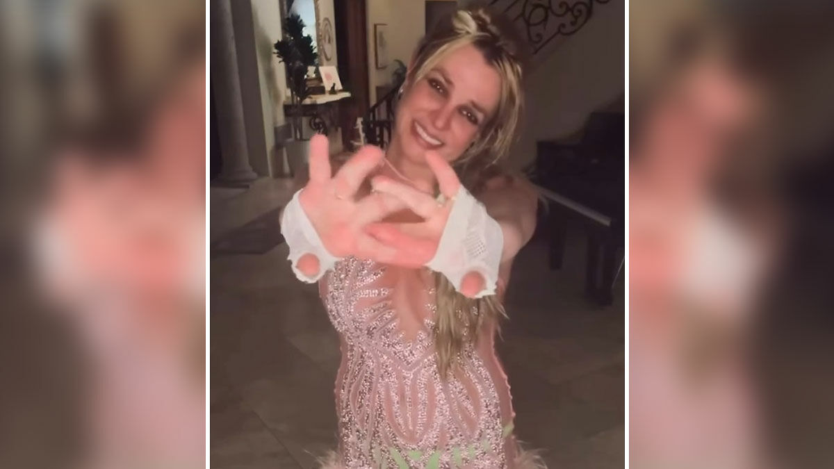 Britney Spears smiling indoors with bruises, wearing a pink dress and fingerless gloves, reaching hands toward the camera.