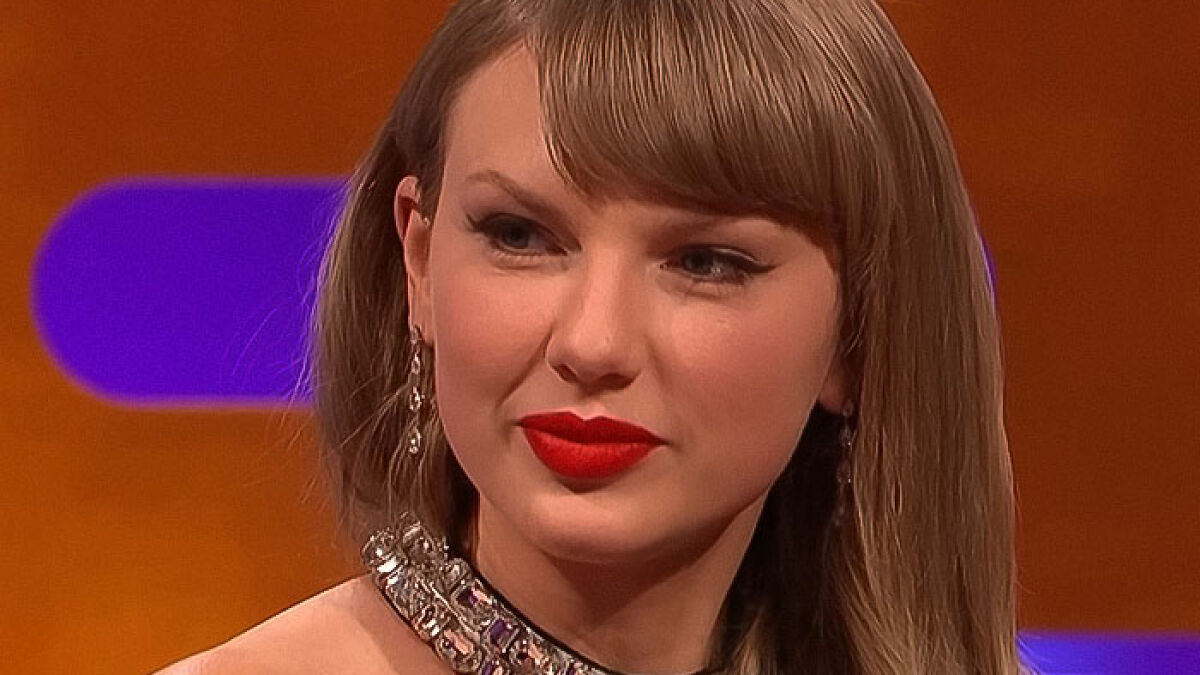 Taylor Swift wearing red lipstick and statement jewelry during an interview on a late-night talk show set.