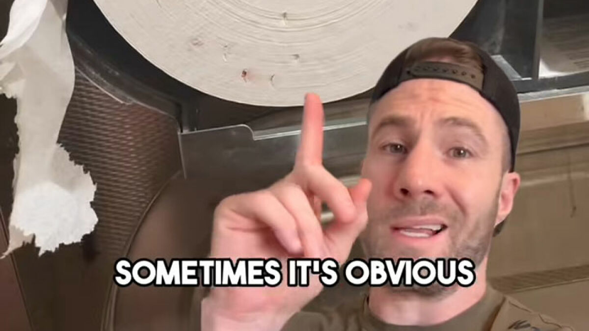 Man wearing a backward cap pointing upward near large toilet paper roll highlighting public bathroom toilet paper danger.