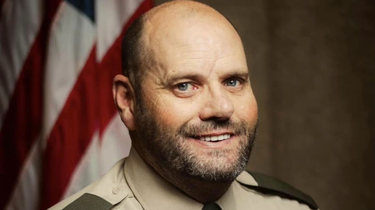 Police chief in uniform smiling slightly with an American flag blurred in the background.