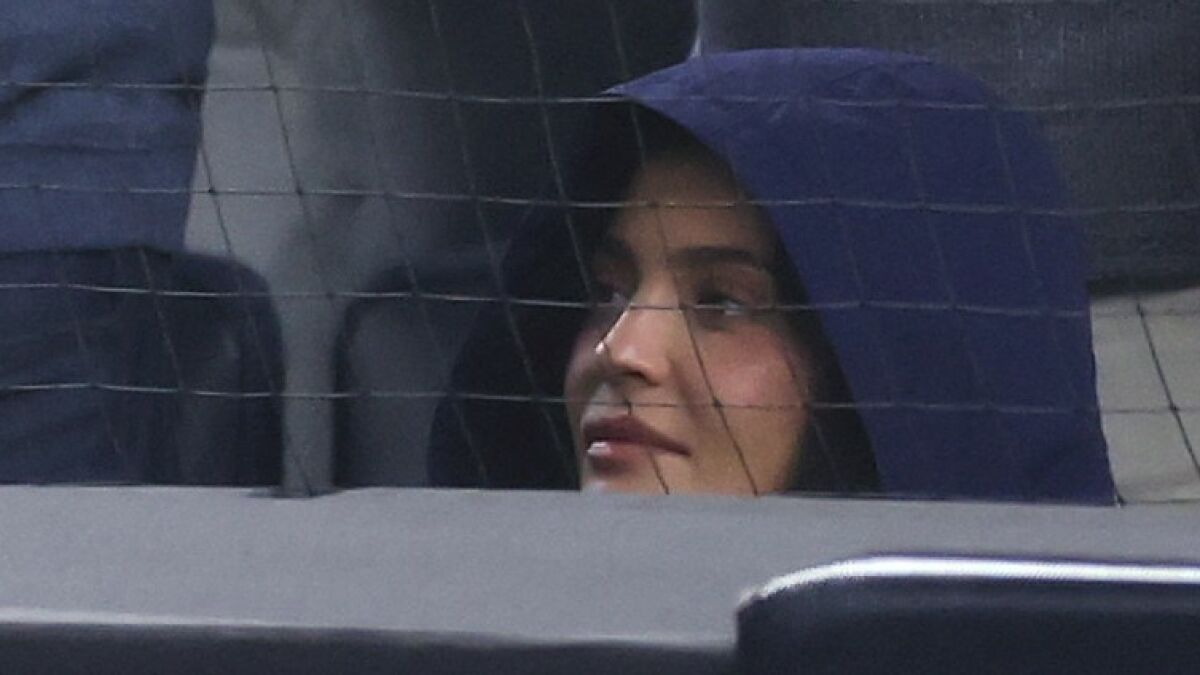 Kylie Jenner wearing a hooded jacket, looking thoughtful and sitting behind a netted barrier.