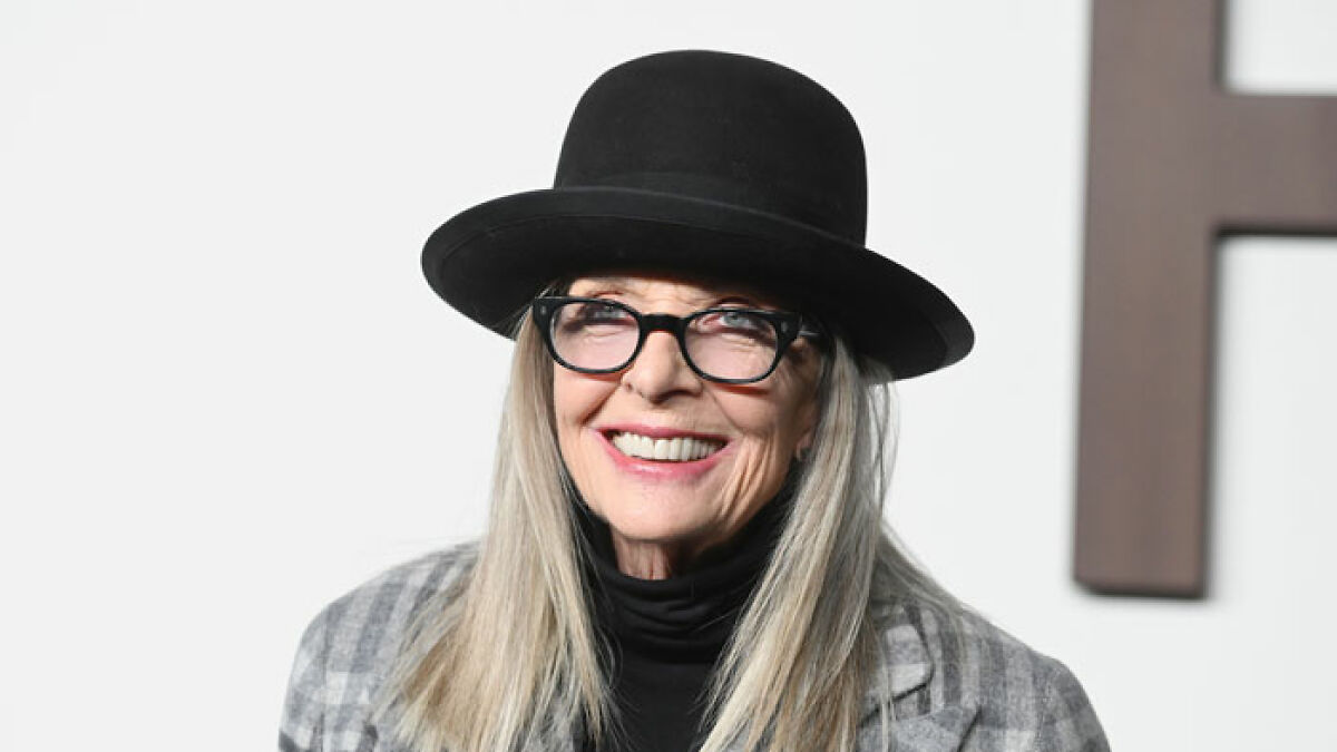 Diane Keaton smiling, wearing a black hat, glasses, and gray checkered jacket, representing Hollywood icon and legend.
