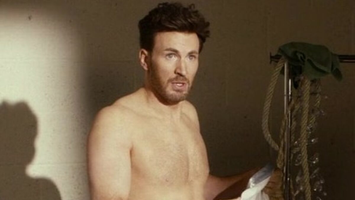 Chris Evans shirtless wearing a jock strap, standing indoors with dim lighting and holding a cloth.