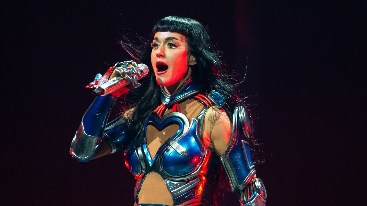 Katy Perry performing on stage, wearing futuristic armor-style costume and holding a microphone during a live concert.