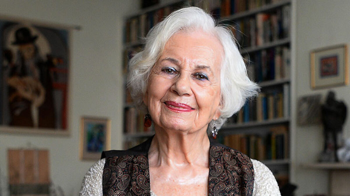 H*******t survivor Ruth Posner smiling softly, wearing a patterned vest, with bookshelves in the background.