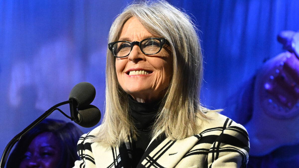 Diane Keaton smiling at a microphone, wearing glasses and a black and white plaid jacket on stage.