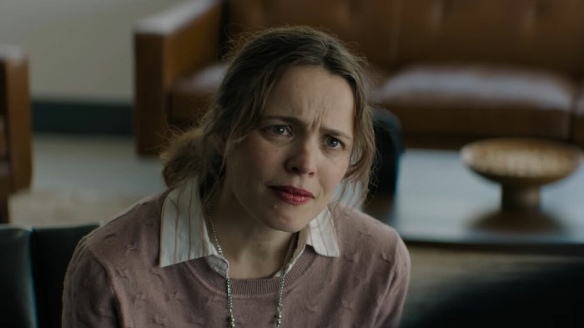 Rachel McAdams with a natural look, wearing a pink sweater and white shirt, appearing emotional in a film scene indoors.