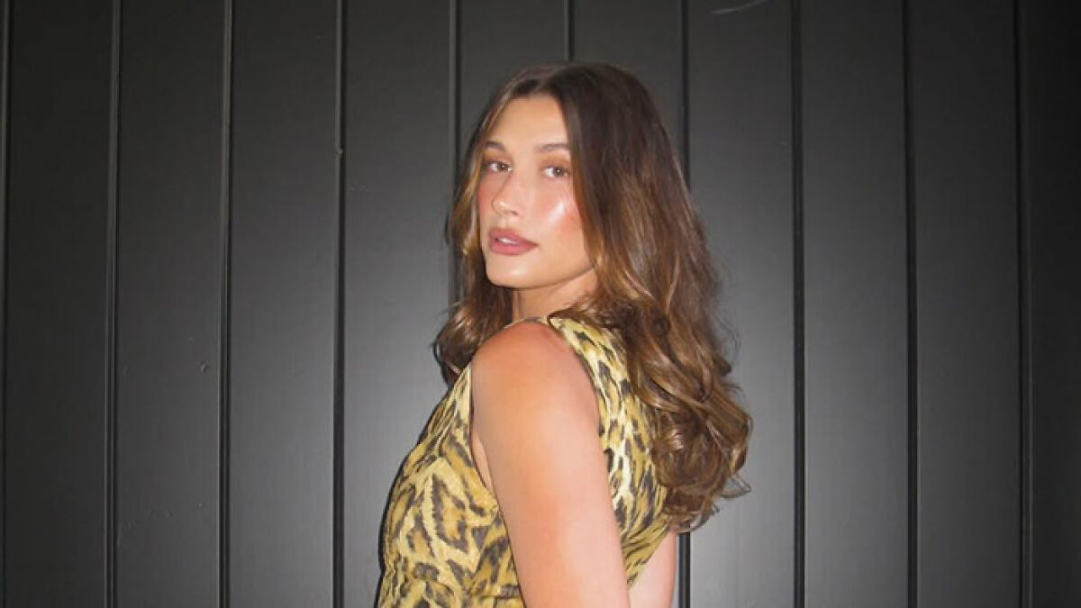 Hailey Bieber posing in a leopard print dress against a dark paneled wall, sparking Selena Gomez feud discussions.