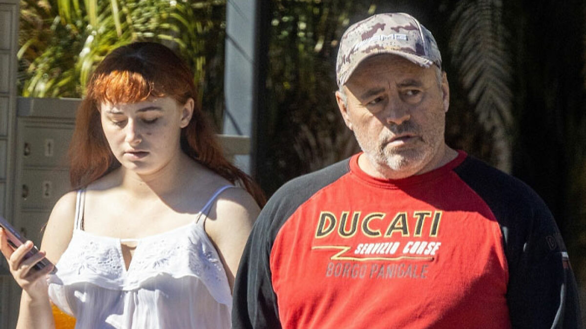 Joe Rogan wearing a red Ducati shirt walking outside with a young woman in a white top, both appearing serious.