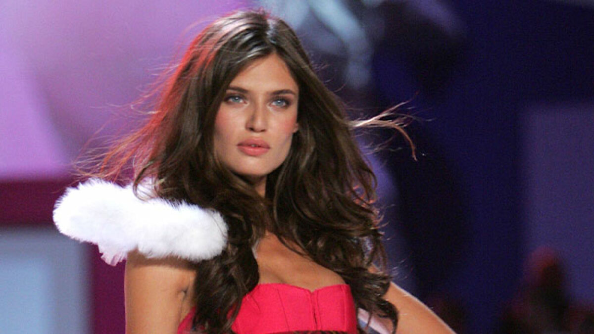 Former Victoriau2019s Secret model with long brown hair wearing a red outfit and white feathered accessory on runway.