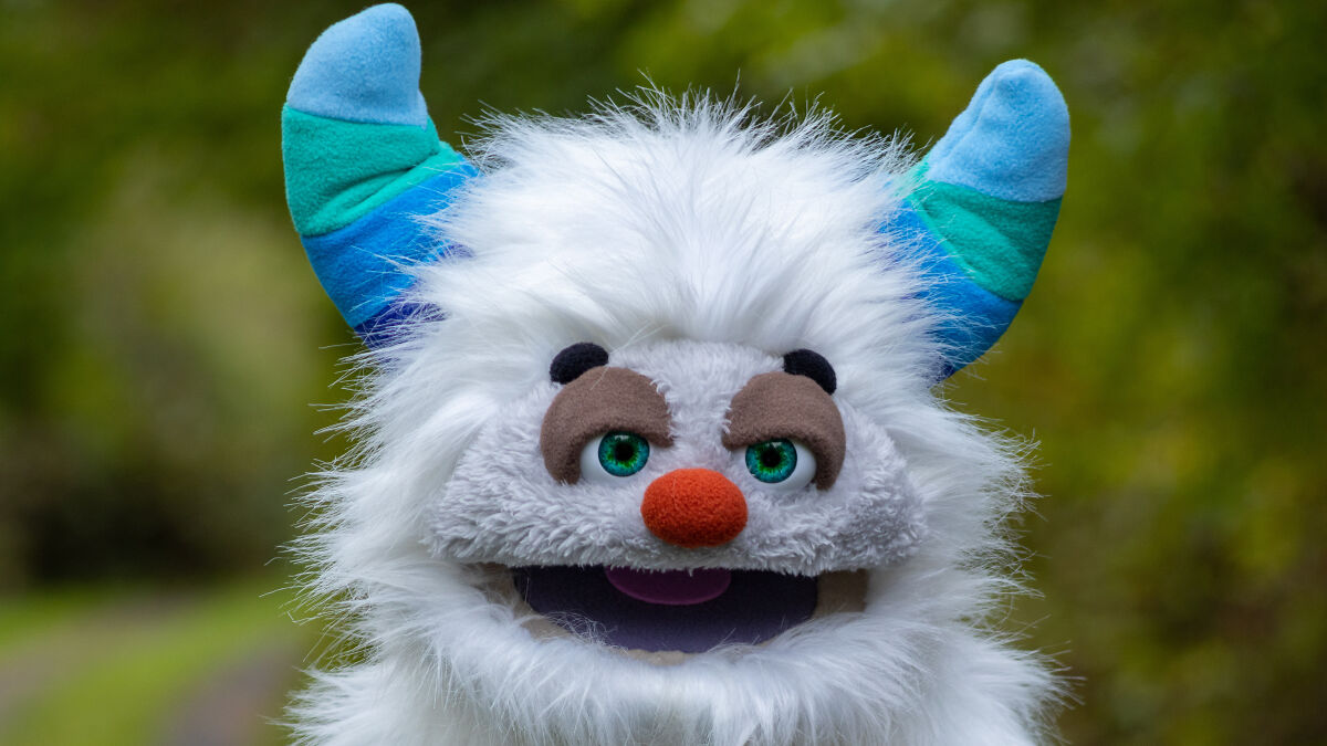A white furry puppet with blue and green striped horns and bright green eyes outdoors for storytelling.