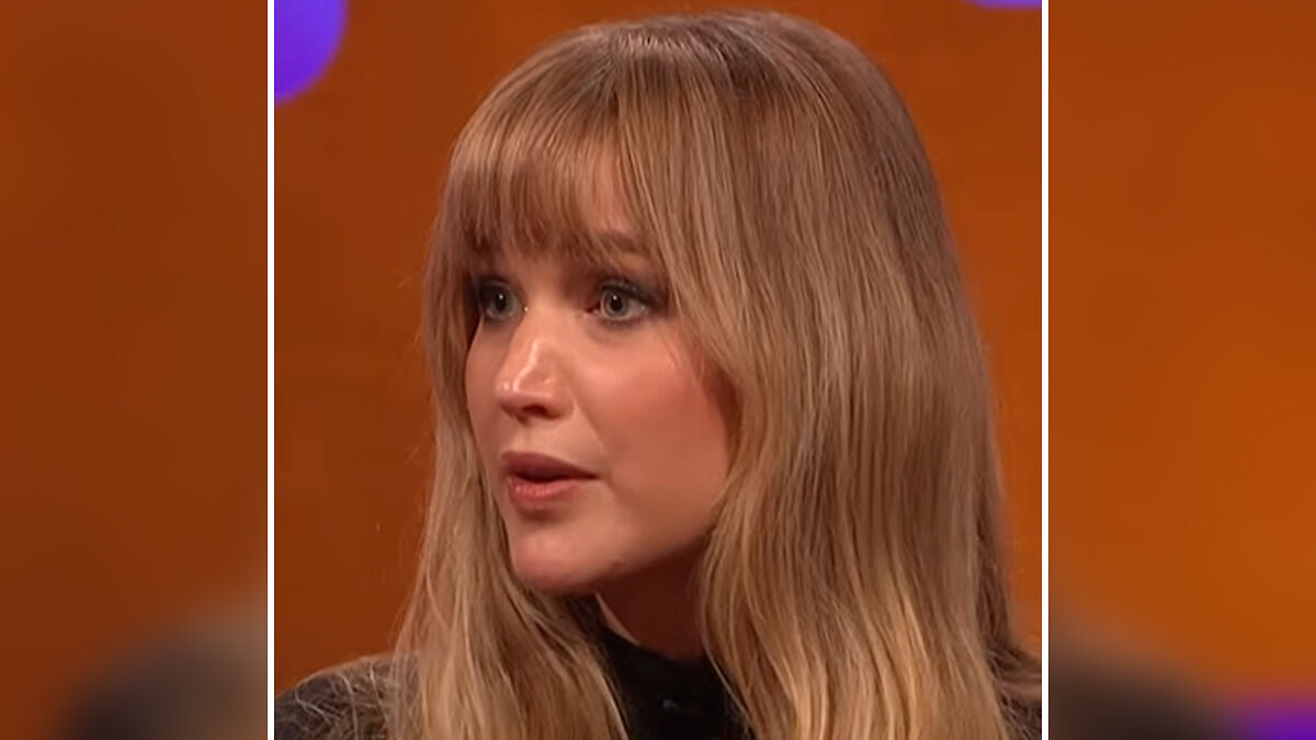 Jennifer Lawrence with long blonde hair responding during a heated moment on a talk show with an orange background.