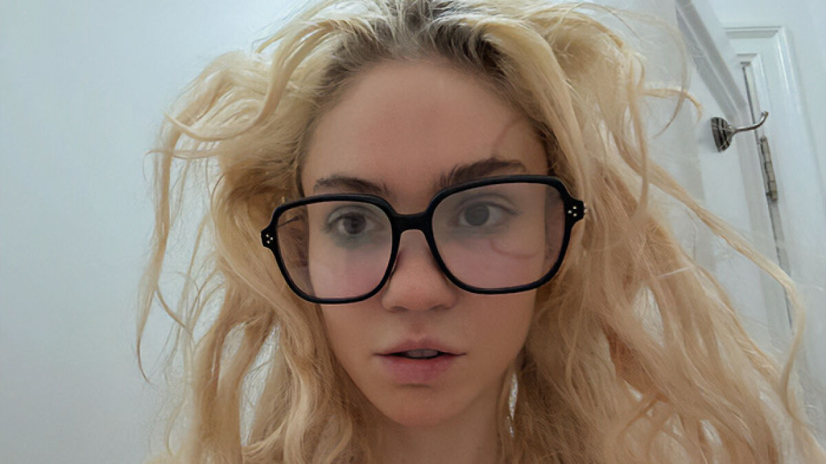Close-up of a woman with blonde hair wearing large black glasses, showcasing her unique face tattoo and style.