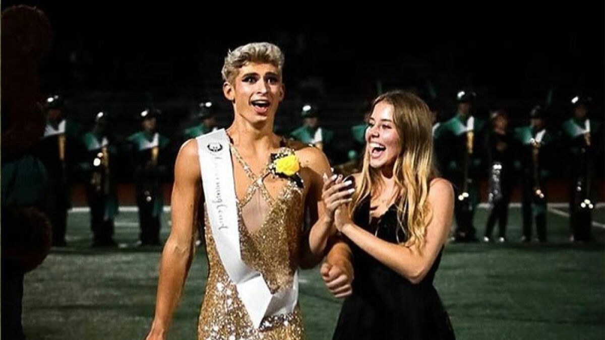 Missouriu2019s first male homecoming queen wearing a gold dress and sash with a smiling woman at a nighttime event.
