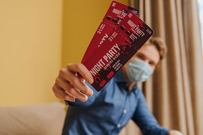 Person wearing mask holding two night party tickets indoors, illustrating coworker show scam realization. Person wearing mask holding two night party tickets indoors, illustrating coworker show scam realization.
