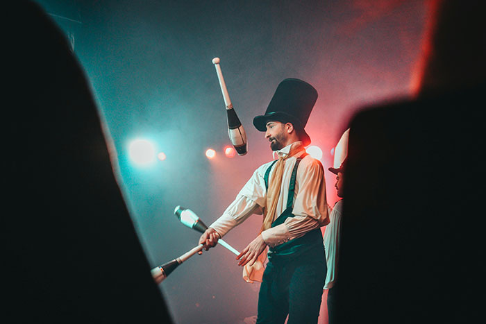 Male performer in a tall hat juggling clubs on stage during a show with dramatic lighting and an audience silhouette. Male performer in a tall hat juggling clubs on stage during a show with dramatic lighting and an audience silhouette.