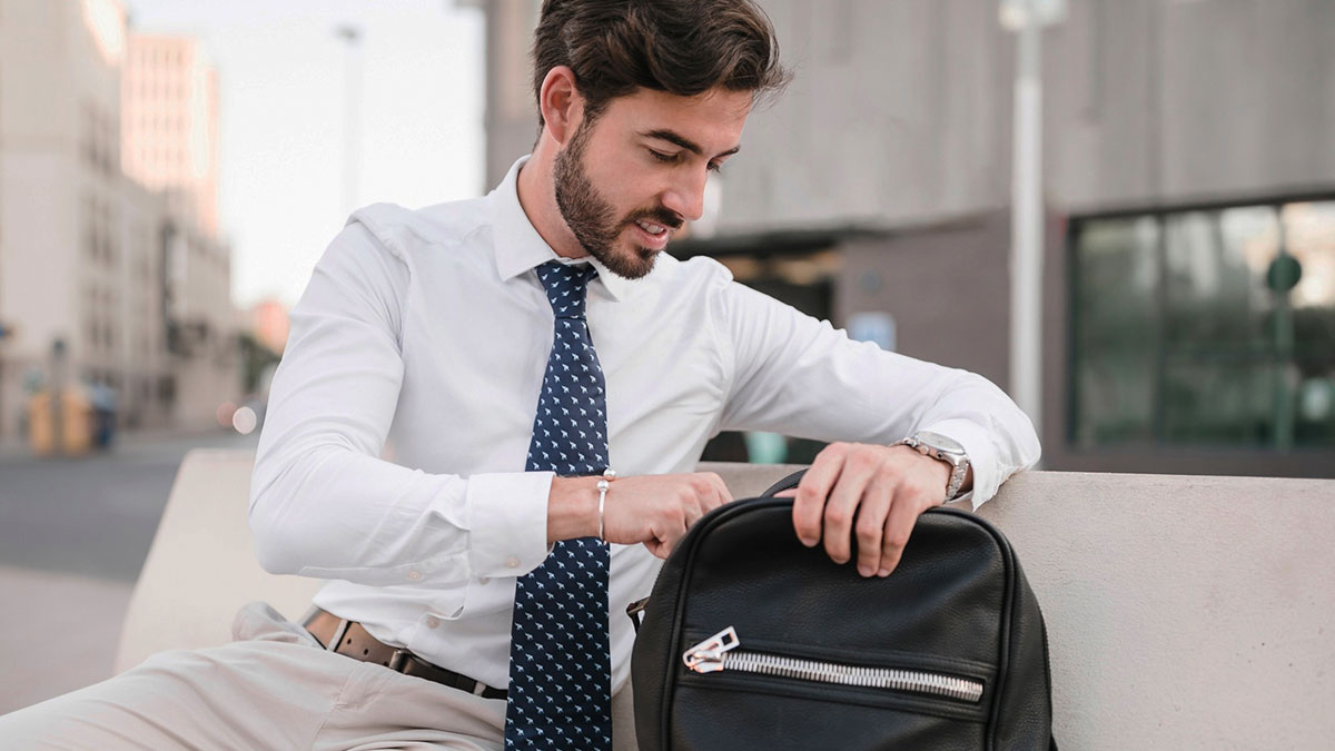 Man in white shirt and tie snooping around in a woman's purse outdoors on a city bench during the day