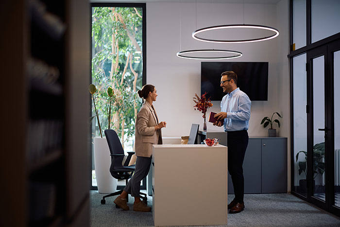Two coworkers in a modern office discussing work nepotism, standing near a desk with plants and circular ceiling lights.