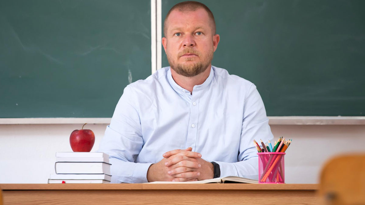 Serious man sitting at a desk with books, an apple, and colored pencils in front of a chalkboard in a classroom setting.