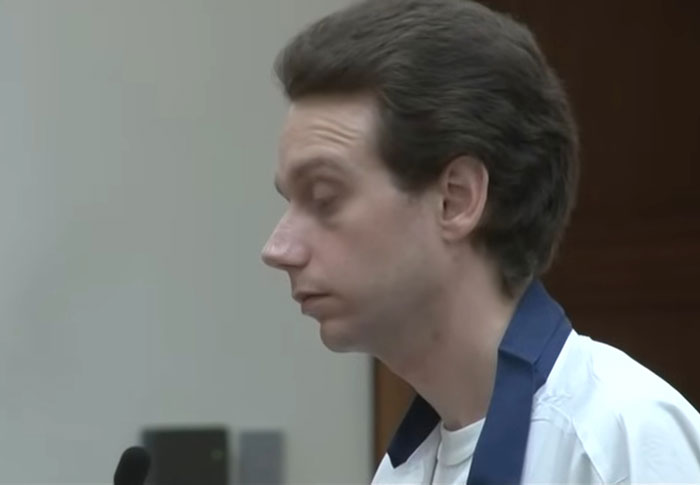 Young man with dark hair and white shirt, standing in a courtroom, reflecting a creepy found footage atmosphere.