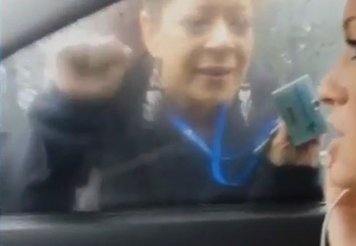 Woman outside a car window holding an object close to another person, evoking a creepy found footage video feel.