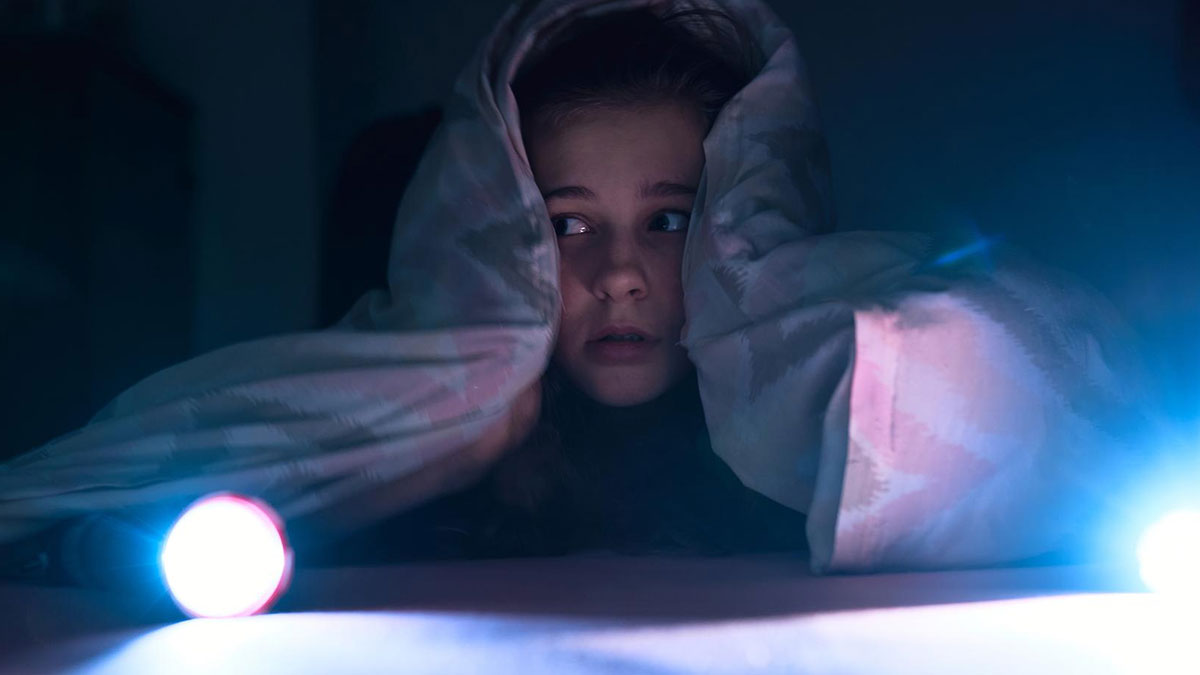 Child hiding under blanket at night with flashlight fearfully looking around in creepy home setting.