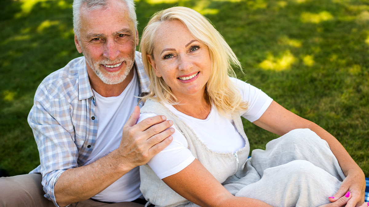 Smiling elderly couple sitting outdoors on grass, capturing a peaceful moment in cozy casual clothing.