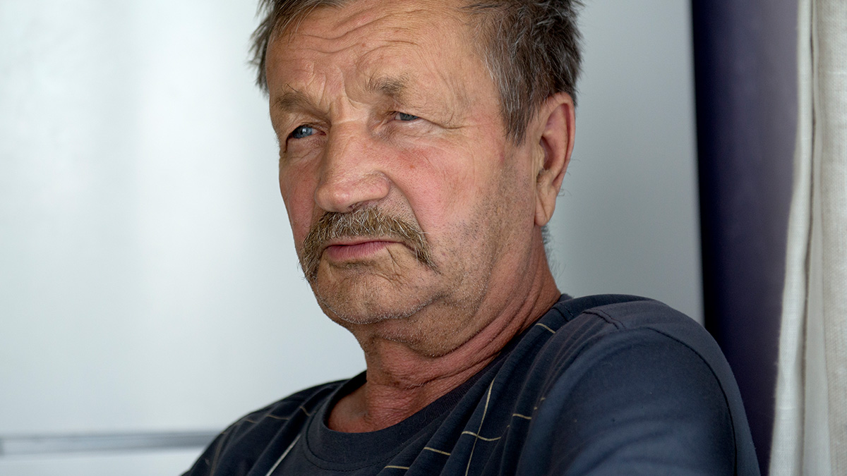 Older man with a mustache looking pensive indoors, reflecting on cult stories from victims or those who knew them