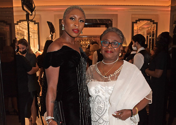 Cynthia Erivo in a black dress and another woman in white, attending an event with guests and photographers in the background. Cynthia Erivo in a black dress and another woman in white, attending an event with guests and photographers in the background.