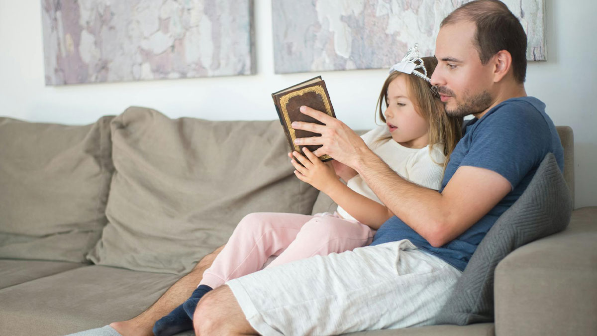 Father reading a book to his daughter on the couch, highlighting the challenge of dating men with kids.