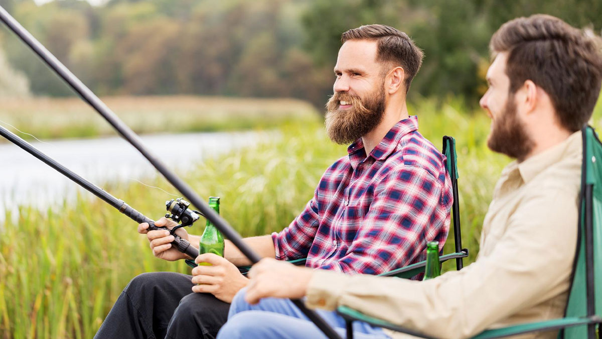 Two men on a boys trip fishing by the lake, sitting with beers and fishing rods during a relaxed outing.