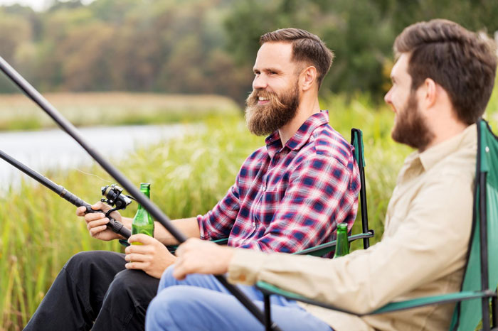 Two men fishing by the lake on a boys trip, one holding a fishing rod and a beer bottle, enjoying outdoors. Two men fishing by the lake on a boys trip, one holding a fishing rod and a beer bottle, enjoying outdoors.