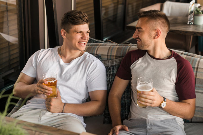 Two men enjoying beers and laughing together on a couch during a casual boys trip outing. Two men enjoying beers and laughing together on a couch during a casual boys trip outing.