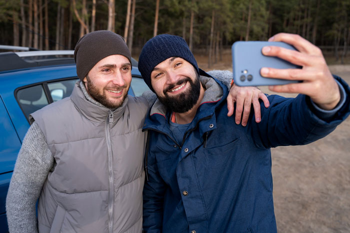 Two men smiling and taking a selfie outdoors during a boys trip in a forested area near a car. Two men smiling and taking a selfie outdoors during a boys trip in a forested area near a car.
