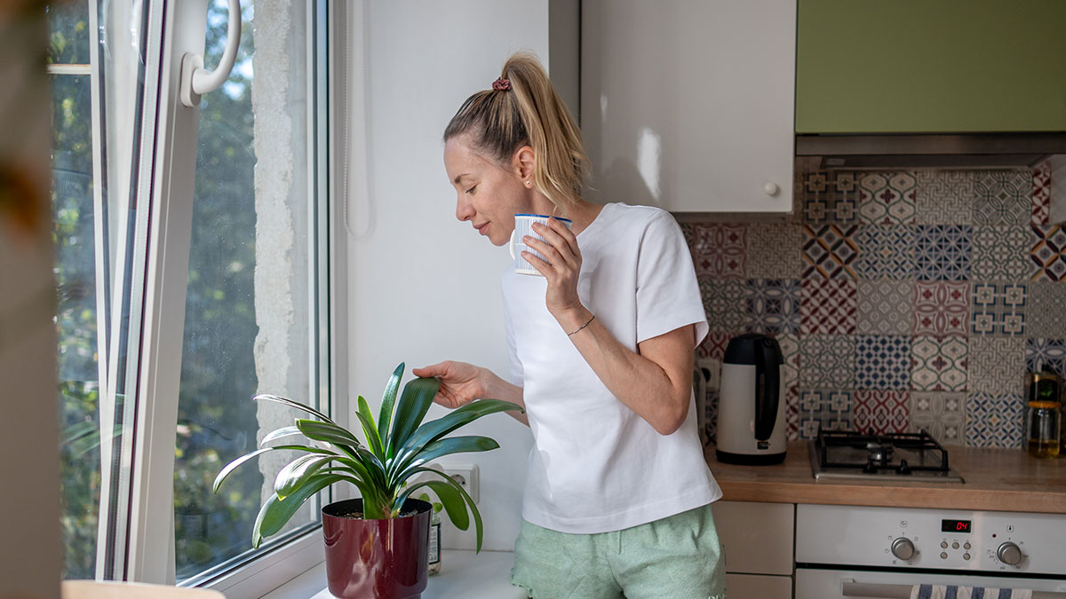 Woman holding a cup in a kitchen, caring for a plant, reflecting on sibling refusing to keep paying rent to parents.