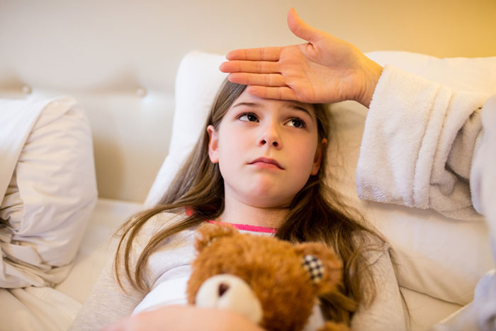 Young girl lying on a couch holding a teddy bear while an exhausted mom checks her forehead for a fever. Young girl lying on a couch holding a teddy bear while an exhausted mom checks her forehead for a fever.