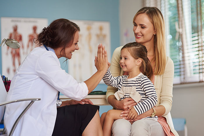 Pediatrician giving high five to a young girl while mother looks on during a child’s medical procedure visit. Pediatrician giving high five to a young girl while mother looks on during a child’s medical procedure visit.