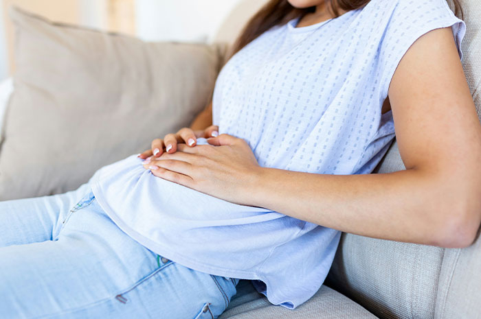 Woman in casual clothes holding stomach in pain on a couch depicting symptoms patients googled before seeing a doctor.