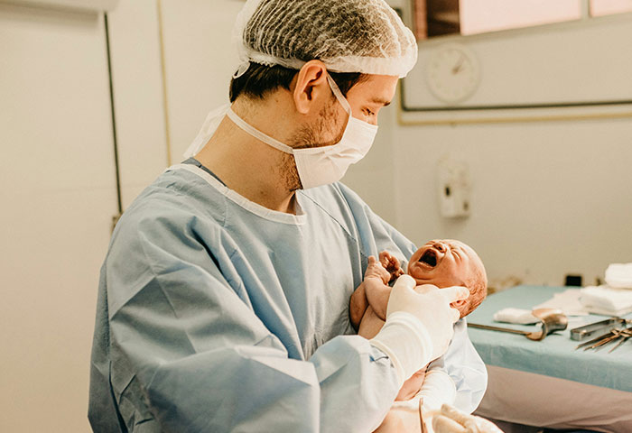 Doctor in protective gear holding a crying newborn baby during birth meltdown witnessed in hospital delivery room