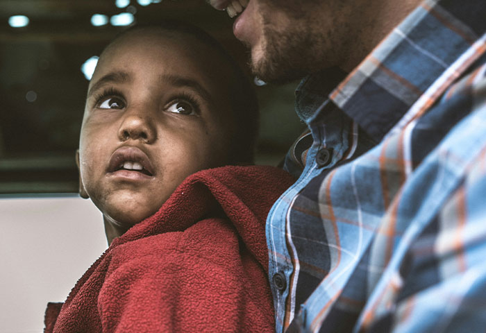 Child in red jacket looking up with concerned expression, sitting next to man in plaid shirt, capturing birth meltdown moment