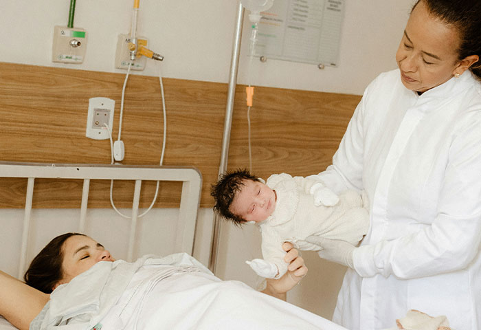 Doctor in white coat holding newborn baby in hospital room while new mother lies in bed after birth meltdown.