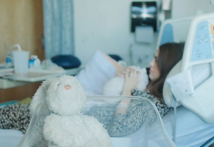 A stuffed animal in a hospital bassinet with a woman resting in a hospital bed during a birth meltdown witnessed by doctors.