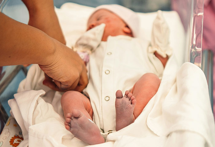 Newborn baby in a white outfit lying in a hospital crib while a hand adjusts the clothing during birth meltdown moments.