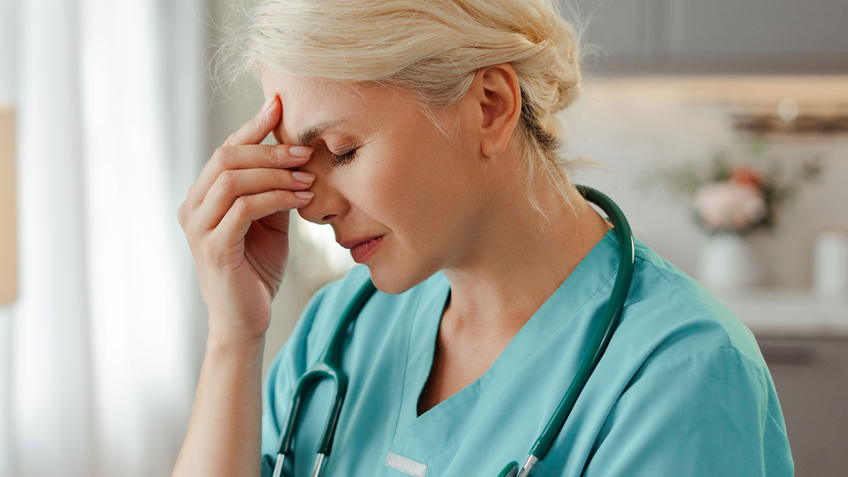 Stressed female doctor in scrubs with a stethoscope, holding her forehead, upset about practice financial loss.