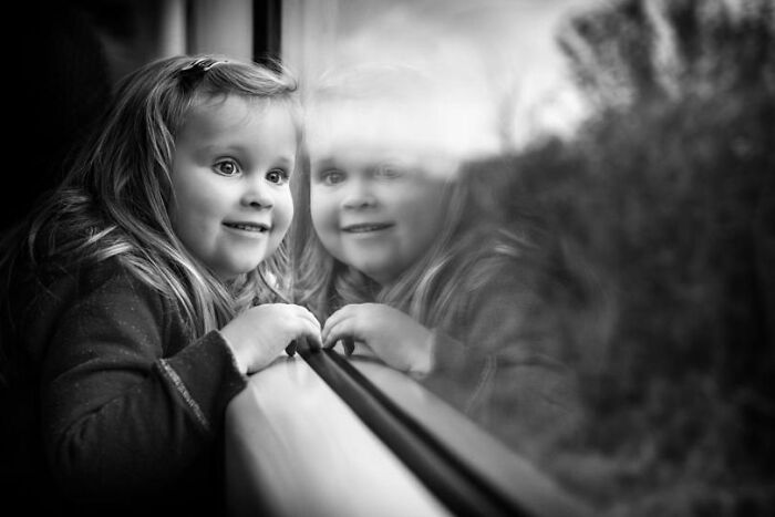 Young girl smiling and looking outside a window, a beautiful moment captured in childhood photography around the world.