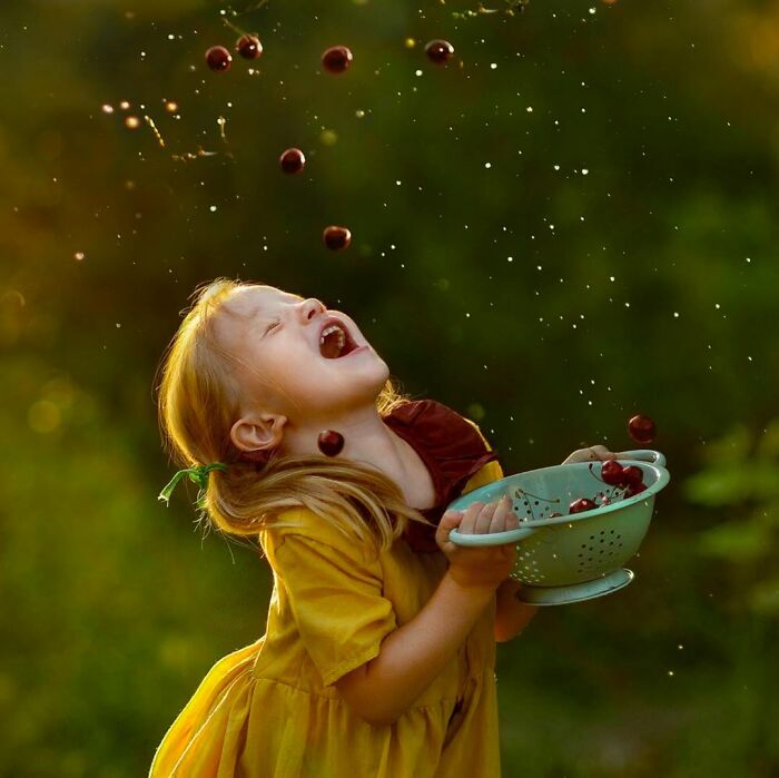 Young girl in a yellow dress joyfully catching cherries in a colander, capturing the beauty of childhood around the world.