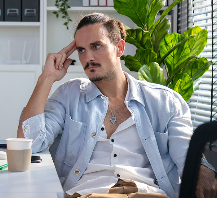 Man wearing light shirt sitting thoughtfully at office desk near plant and window blinds, easily replaceable employee concept. Man wearing light shirt sitting thoughtfully at office desk near plant and window blinds, easily replaceable employee concept.