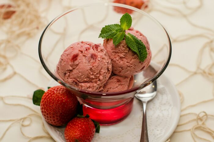 Strawberry ice cream scoops garnished with mint in a glass bowl alongside fresh strawberries on a white plate.