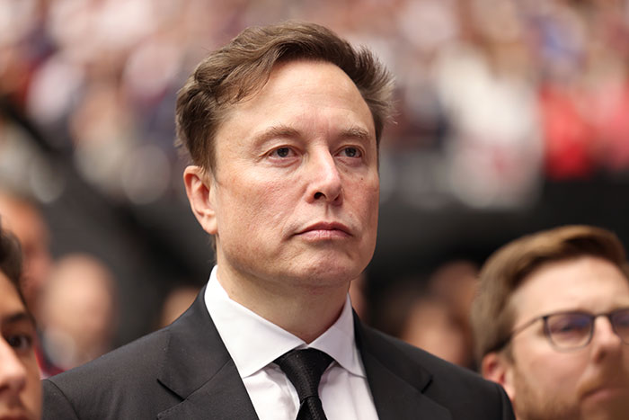 Elon Musk at a public event, wearing a black suit and tie, with blurred crowd in the background.